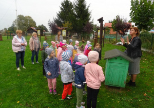 Grupa dzieci zebrana jest wokół ula razem z panią dyrektor, panią Agnieszką i panią Anią. Słuchają opowiadającej z zaangażowaniem pani w czarnej kurtce na temat produkcji miodu.